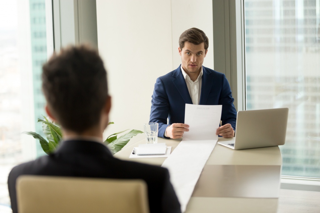 Employer sits across a long table from an interviewee. He holds one end of an extremely long paper that begins at the opposite end of the table.