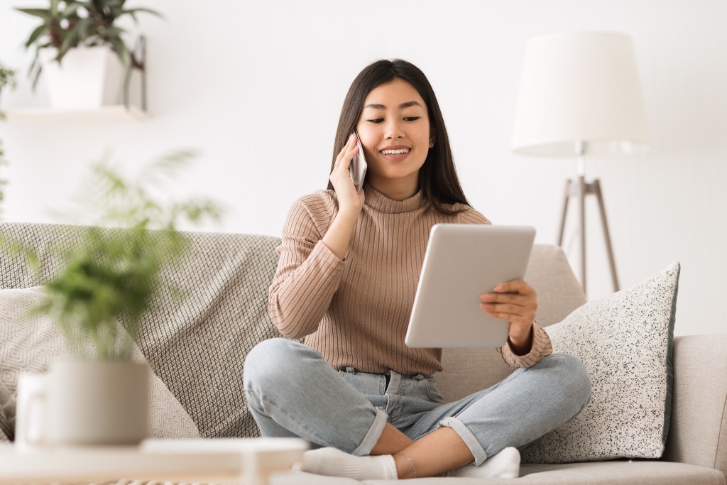 Young woman sits cross-legged on a sofa, smiling as she looks at a large pad and speaks on her cellphone. 