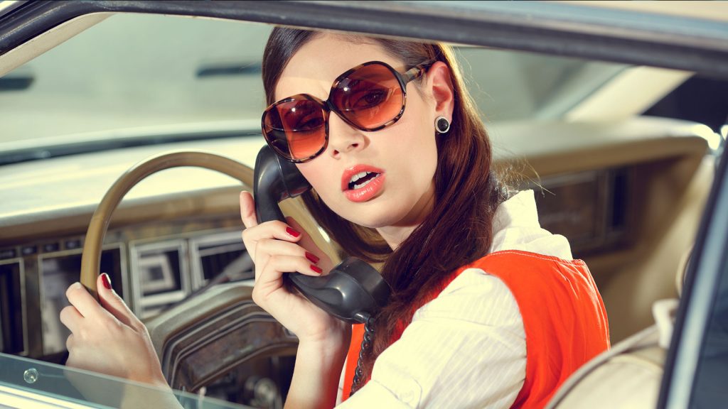 A distracted female driver, vintage '60's style, in tinted sunglasses and bright lipstick speaks on a carphone while looking backwards.
