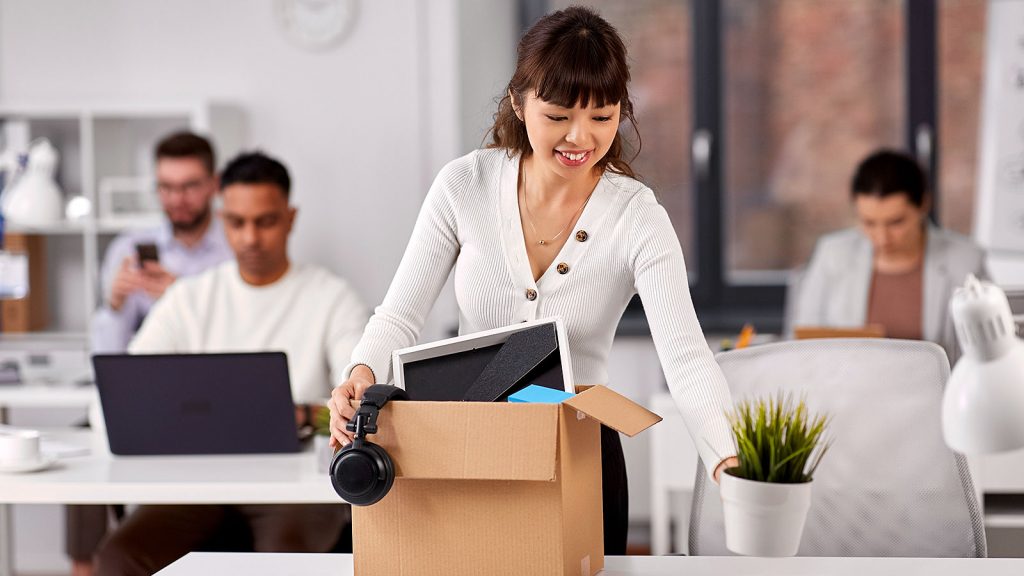 A happy female packs her desk belongings into a cardboard box after quitting her job.