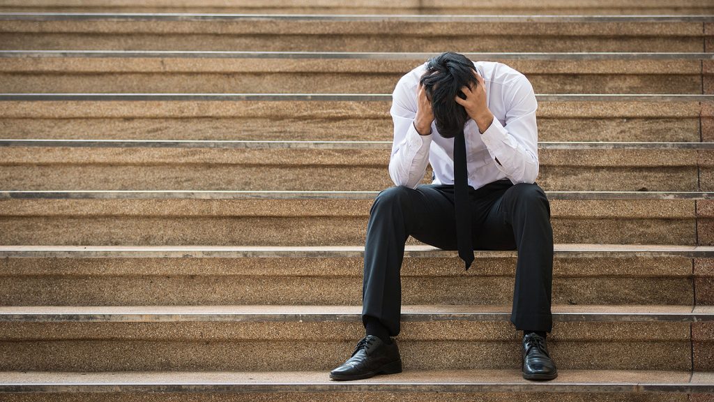 An unhappy male office worker sits outside on steps and holds his head in his hands.